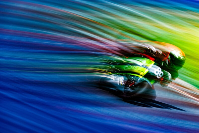 A motorcycle racer in green gear speeds through a track, creating colorful motion blur streaks in blue, red, and yellow tones, emphasizing the sense of rapid movement and vibrant energy.