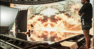A person floats in a shallow pool on a film set, surrounded by equipment. A large digital backdrop displays a dramatic cloud scene, while another person stands nearby, observing the setup.