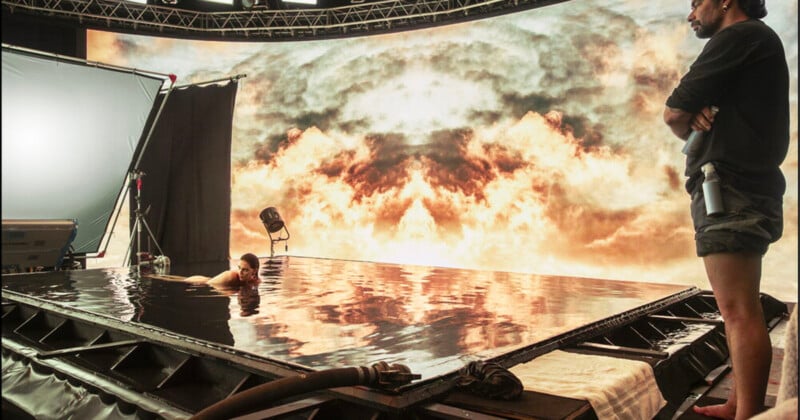 A person floats in a shallow pool on a film set, surrounded by equipment. A large digital backdrop displays a dramatic cloud scene, while another person stands nearby, observing the setup.