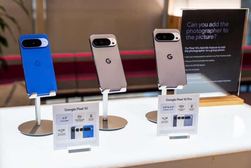 Three Google Pixel smartphones are displayed on stands with info cards beneath them. A sign in the background describes a photo feature. The phones are in blue, gray, and black colors.