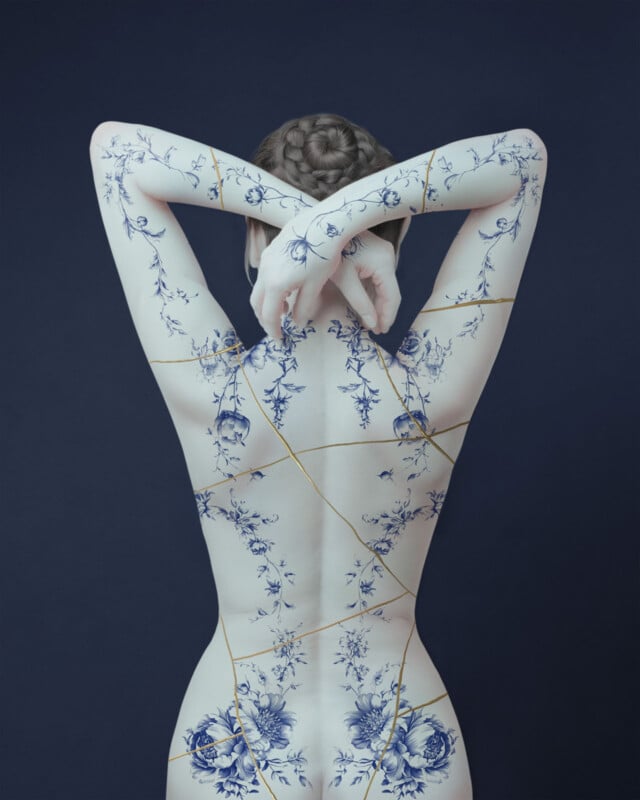 A person with braided hair poses with arms raised, showing their back covered in blue floral porcelain patterns and gold cracks, resembling kintsugi on a dark blue background.