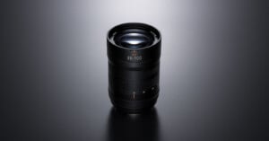 A black camera lens with “35–100” marking is standing upright on a smooth, dark surface under soft lighting, highlighting its textured grip and glass elements.