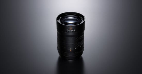 A black camera lens with “35–100” marking is standing upright on a smooth, dark surface under soft lighting, highlighting its textured grip and glass elements.