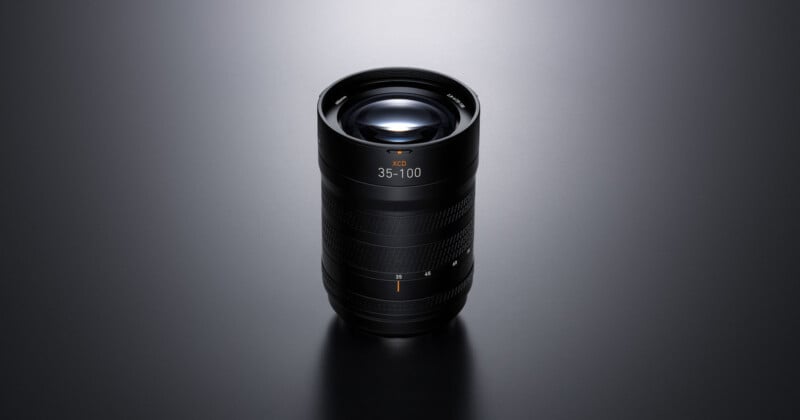 A black camera lens with “35–100” marking is standing upright on a smooth, dark surface under soft lighting, highlighting its textured grip and glass elements.