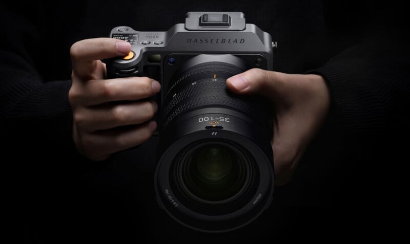A person in a dark sweater holds a large Hasselblad camera with both hands, focusing on the camera's lens and controls against a black background.