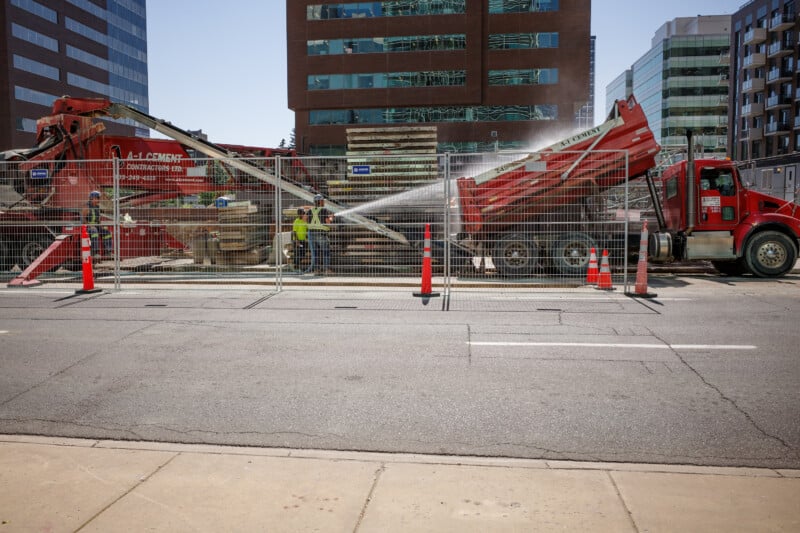 Construction https://creatorkitdepot.com/ on a city street with workers, orange cones, a red cement truck, and a water hose spraying over materials. Office buildings are visible in the background behind a metal fence.