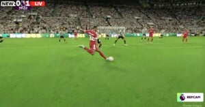A Liverpool player in a red kit kicks a soccer ball during a match against Newcastle United, with the goalkeeper and defenders ready. The score is 0-1, and the crowd fills the stadium seats.