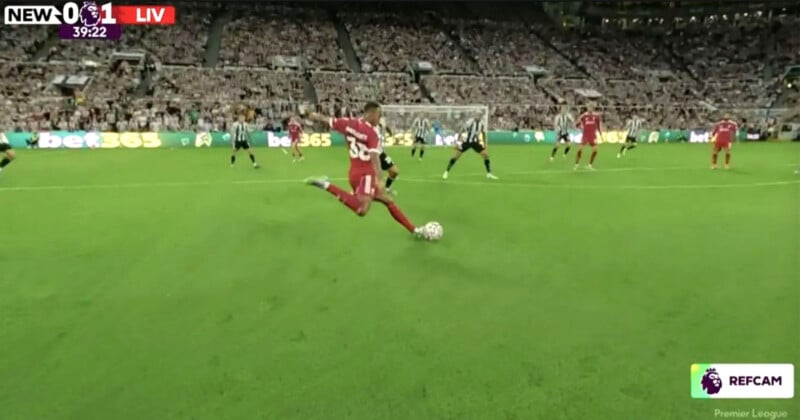A Liverpool player in a red kit kicks a soccer ball during a match against Newcastle United, with the goalkeeper and defenders ready. The score is 0-1, and the crowd fills the stadium seats.