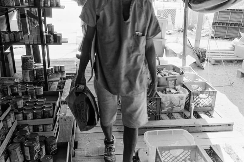 A person in casual clothes carries a bag through a makeshift pantry stocked with canned goods and food crates, with shelves and boxes organized on wooden pallets.