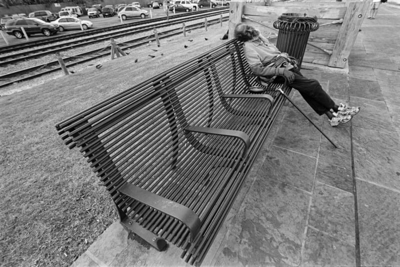 A person lies sprawled on a metal bench near train tracks, appearing to rest with their head back and one arm over their face. Cars are parked in the background, and a trash can sits nearby on a stone-paved area.