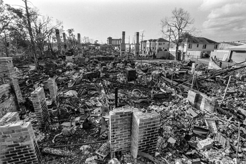 Black and white image of a neighborhood devastated by fire, with charred debris, collapsed structures, and standing brick chimneys. Nearby intact houses remain, and leafless trees are visible against a cloudy sky.