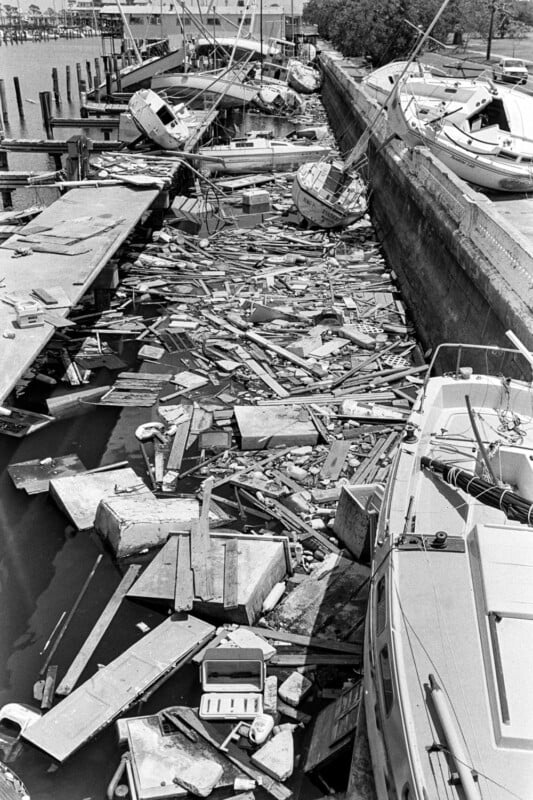 A dock and boats heavily damaged by a storm; debris, wood planks, and broken parts fill the water along the pier, with several tilted and wrecked sailboats lining the edge.