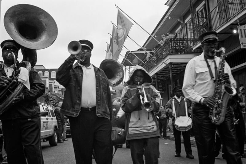 Black and white photo of a street band playing brass instruments while marching; people in uniforms and hats lead, with buildings, flags, and a crowd in the background.