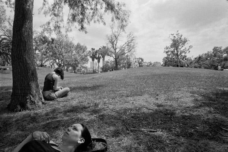 A woman lays on the grass in the foreground while another sits under a tree in the background on a sunny day in a park with scattered trees and a gently sloping hill.