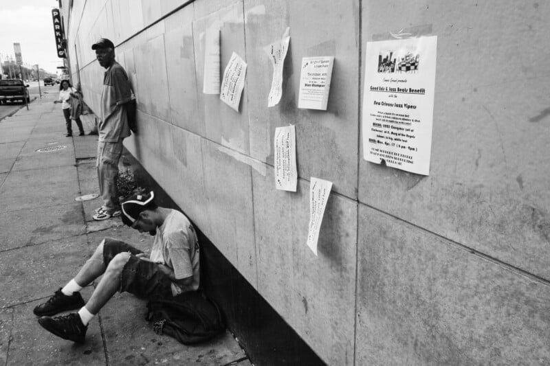 Two men on a city sidewalk next to a wall with torn and weathered flyers; one man sits on the ground looking down, while the other stands leaning against the wall.