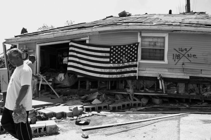 A damaged house with an upside-down American flag hanging from the front, debris scattered around, and two men in front—one walking away and another inside the open doorway. Spray-painted markings are visible on the house.