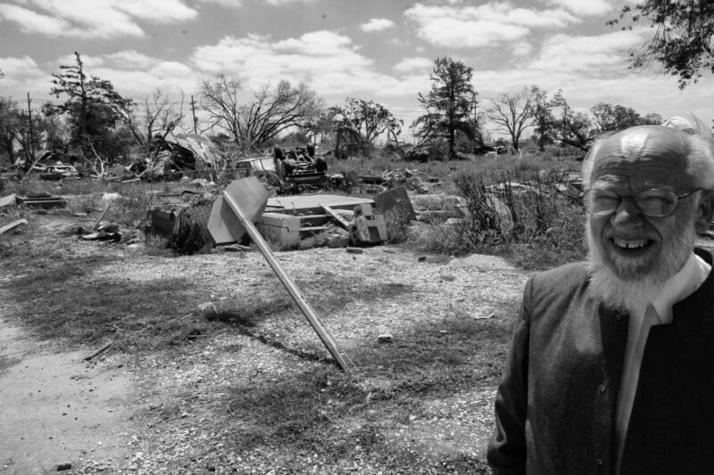 An elderly man with glasses and a beard stands smiling in the foreground, while behind him is a scene of devastation with debris, a fallen sign, and damaged trees under a partly cloudy sky.