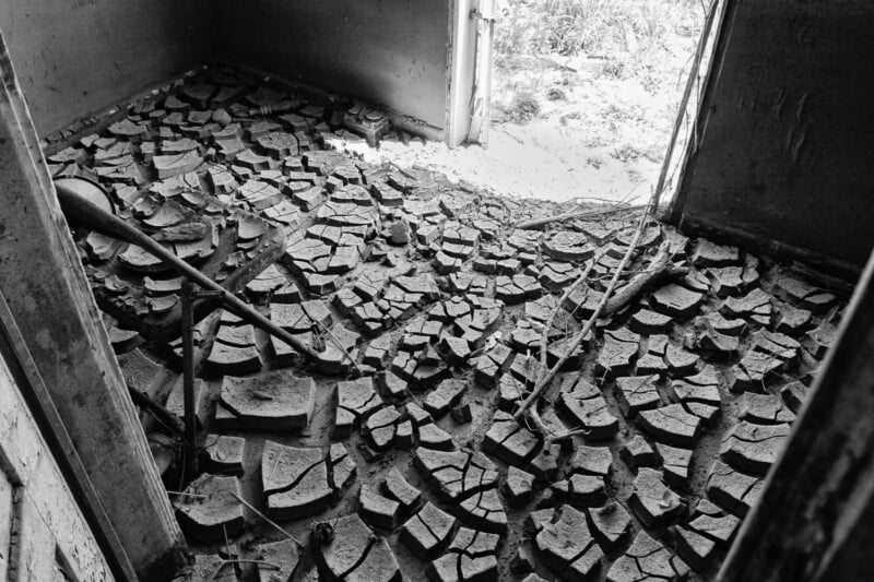 A black and white photo of a room with a cracked, dry mud floor. Pieces of the floor are broken and uneven. The room appears abandoned, and sunlight enters through a doorway leading outside.