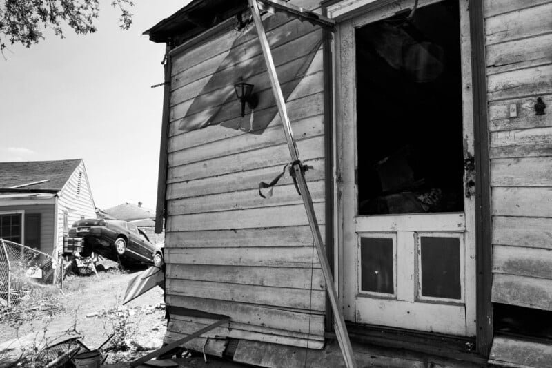 A damaged wooden house with broken siding and an open door. In the background, a car is tilted on top of debris near another house. The scene appears chaotic and shows signs of destruction.