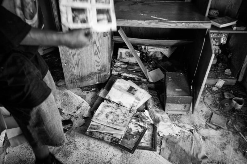 A black and white photo showing a person holding damaged photos, with more water-damaged photographs, debris, and broken furniture scattered on the floor of a messy, abandoned room.