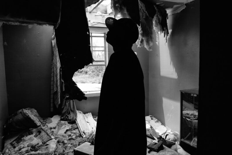 A person in silhouette looks up at a large hole in the ceiling of a damaged room filled with debris, torn insulation, and scattered papers, with daylight shining through the opening.
