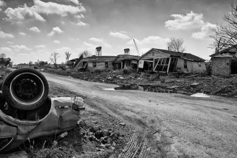 An overturned car lies on a muddy roadside in front of damaged, abandoned houses. The area appears desolate, with debris scattered, broken windows, and a cloudy sky overhead.