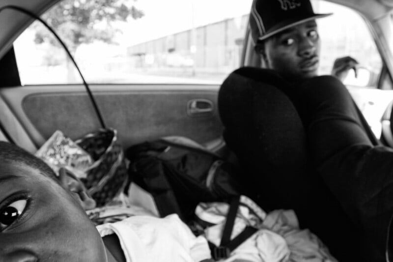 A black-and-white photo showing a close-up of a child’s face in the foreground and a man in a baseball cap looking back from the driver’s seat in a cluttered car interior.