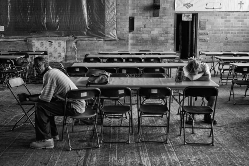Two people sit at opposite ends of a long table in a large, empty room. One looks into the distance, while the other rests their head on folded arms, suggesting fatigue or sadness. Several empty chairs surround them.