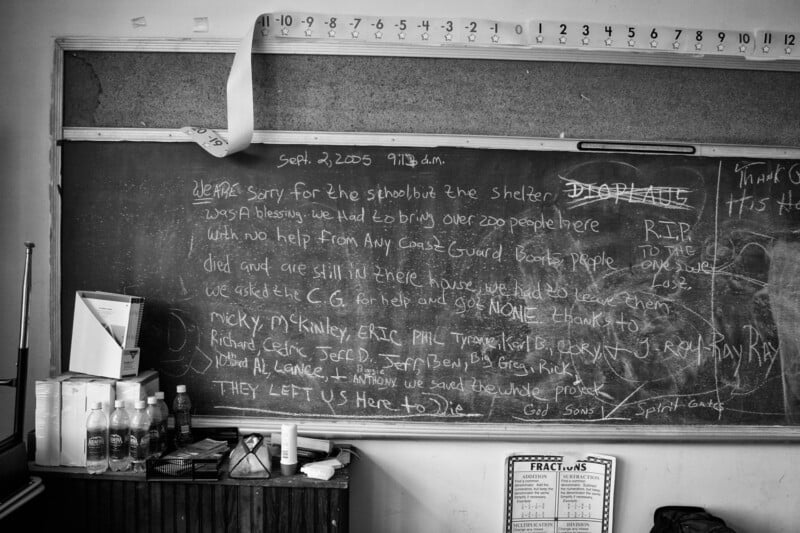 A black-and-white photo of a classroom chalkboard with handwritten messages about being left in a shelter after Hurricane Katrina. Bottles and other items sit on a shelf below the board; a number line and fraction chart are visible.