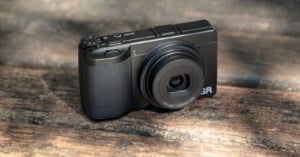 A compact black digital camera with a textured grip and extended lens sits on a wooden surface in natural light.