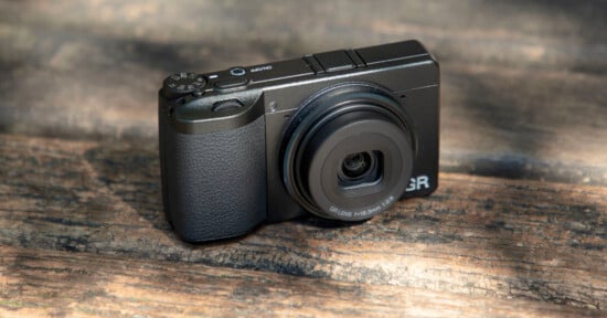 A compact black digital camera with a textured grip and extended lens sits on a wooden surface in natural light.