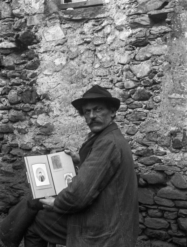 A man in a hat and coat stands in front of a rough stone wall, holding an open photo album with portraits visible, and looking seriously at the camera.