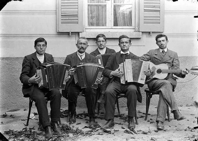 Five men sit in a row outdoors, four holding accordions and one with a guitar. They wear suits and pose in front of a building with a window and open shutters. Dry leaves are scattered on the ground.