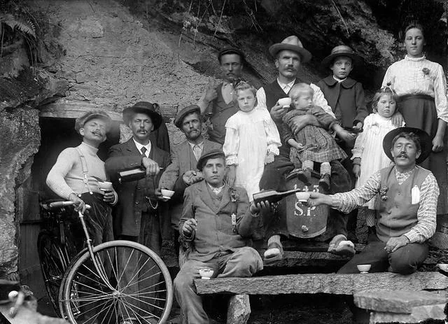 A black-and-white photo of a group of men, women, and children posing outdoors with cups, a bicycle, and a wooden barrel. Some are seated on stone steps and ledges, while others stand behind them, dressed in early 20th-century clothing.