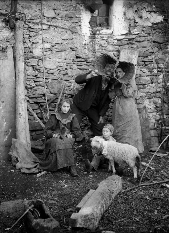 A black and white photo of four people in old-fashioned clothing posing by a stone wall: two adults stand with a tool, two children sit on the ground, and a sheep stands near them. The scene looks rustic and historical.