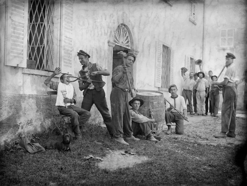 A group of men in work clothes stand and sit outside a building, some holding drinks and tools. A few are raising their hats or posing playfully. There is a barrel nearby and a small dog resting on the grass.