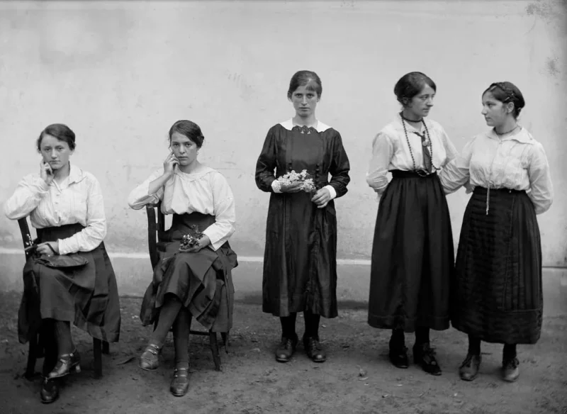 Five young women, dressed in early 20th-century clothing, pose for a black-and-white photo. Three are seated with flowers in their laps, while two stand together, one holding flowers, against a plain wall background.