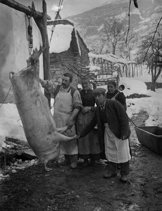 Four people in winter clothing stand outdoors in snow next to a large pig hanging upside down from a wooden frame. Old buildings, bare trees, and mountains are visible in the background. The scene appears historical and rural.