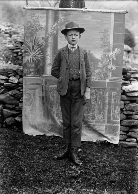 A boy in early 20th-century attire, including a suit, tie, and hat, stands on grass in front of a stone wall and a painted backdrop with plants and columns. His expression is serious and his hands are in his pockets.