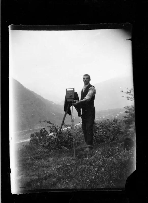 A man stands outdoors next to a large, old-fashioned camera on a tripod, with hills and a river visible in the background. He looks toward the camera, surrounded by grass and foliage. The photo is in black and white.
