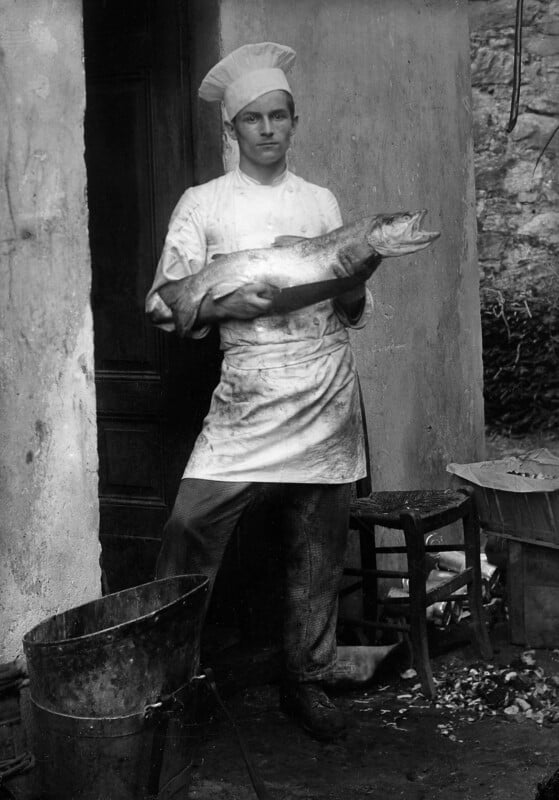 A chef in a white hat and apron stands outside holding a large fish. He appears serious and is standing near a wooden door, with buckets and chairs around him on a stone surface.