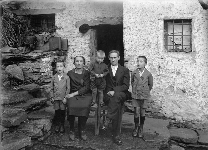 A black-and-white photo of a family of five—two adults and three boys—posing outside a rustic stone house. The adults are seated, the youngest child sits on their lap, and the two older boys stand on either side.