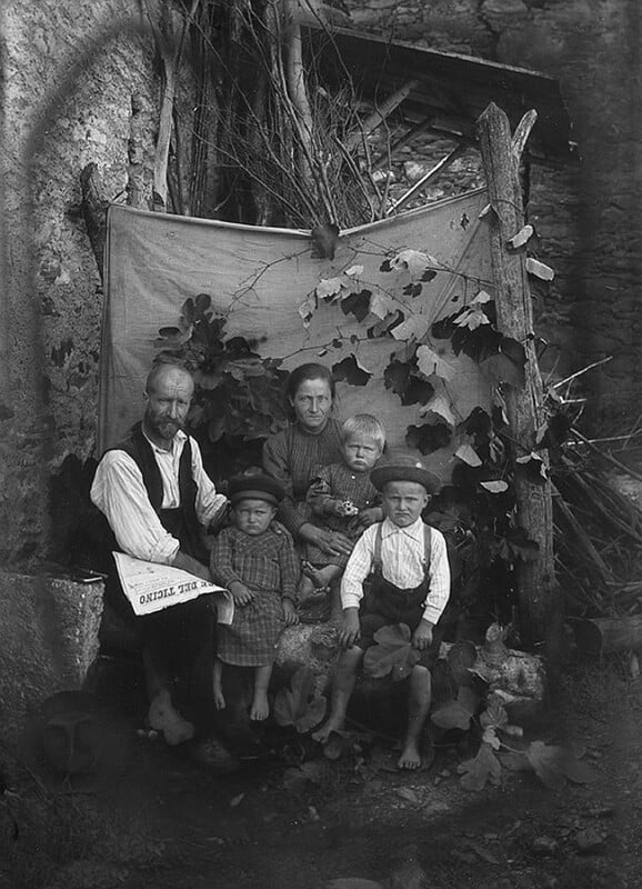 A black and white photo of a family—two adults and three children—posing barefoot in front of a rustic backdrop with branches and leaves, wearing old-fashioned clothing and somber expressions.