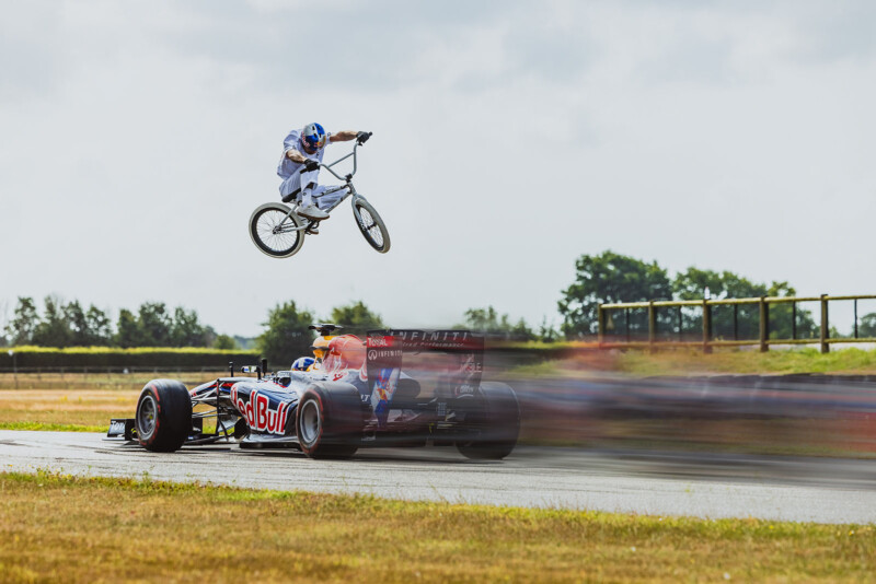 A BMX rider performs a high jump over a speeding Red Bull Formula 1 car on a racetrack, with motion blur showing the car's fast movement and a grassy field in the background.