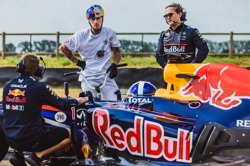 Four people are gathered around a Red Bull Formula 1 car on a racetrack. Two wear Red Bull team gear, one in a white shirt, and one person sits in the car. They appear to be discussing or preparing for an event.