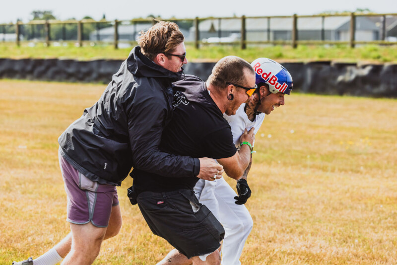 Two men enthusiastically hug or tackle a man wearing a Red Bull helmet and white suit on a grassy field, all appearing to be having fun outdoors.