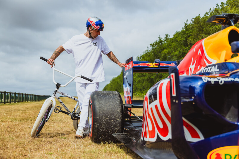 A man in white clothes and a Red Bull helmet stands next to a BMX bike, touching the rear wing of a Red Bull Formula 1 car parked on grass, with trees and a cloudy sky in the background.