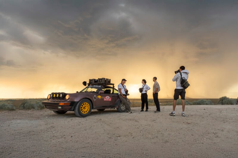 Four people stand beside a modified brown car with yellow wheels on a dirt road, while one person takes photos. The sky is dramatic with a golden hue and a storm or rain visible in the distance.