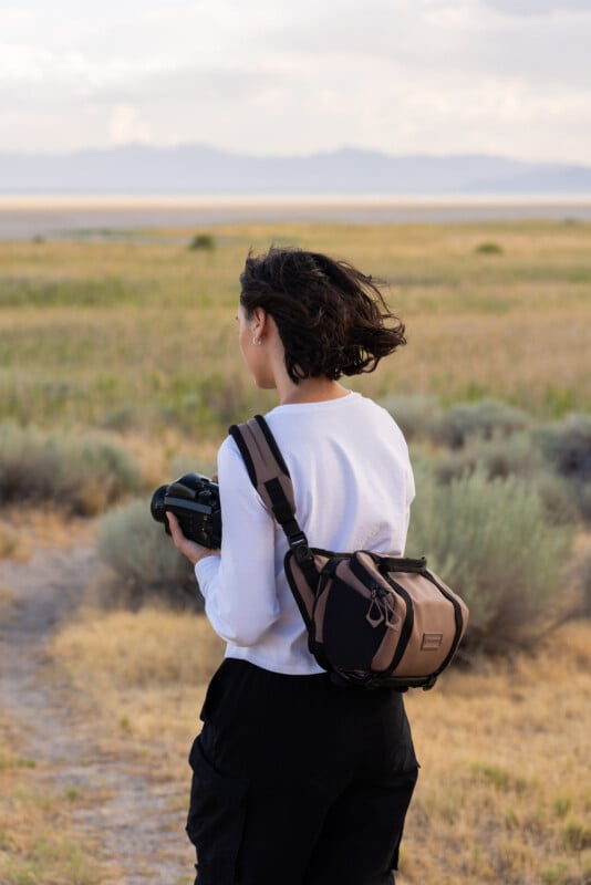 A person with short dark hair, wearing a white long-sleeve shirt and black pants, holds a camera while standing in a grassy, open landscape with mountains in the distance. Their back is facing the camera.