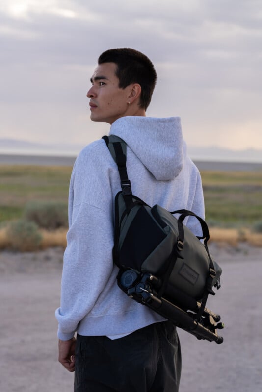 A young man in a gray hoodie looks over his shoulder while standing outdoors. He carries a black camera bag with a tripod attached, with a landscape of grass, dirt, and distant mountains behind him.
